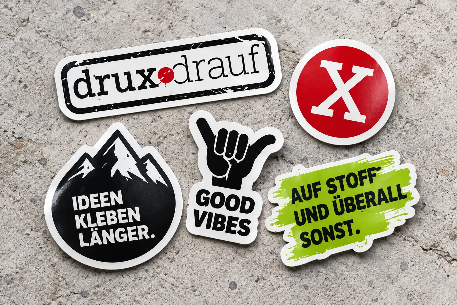 Sticker-Mockups auf Beton: DRUXDRAUF-Logo, rotes X, Bergmotiv, Good Vibes und grüner Schriftsticker mit weißen Konturschnittkanten